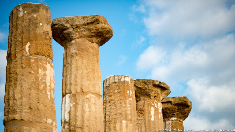 Upward view of several weathered Doric columns with broad capitals against a blue and cloudy sky.