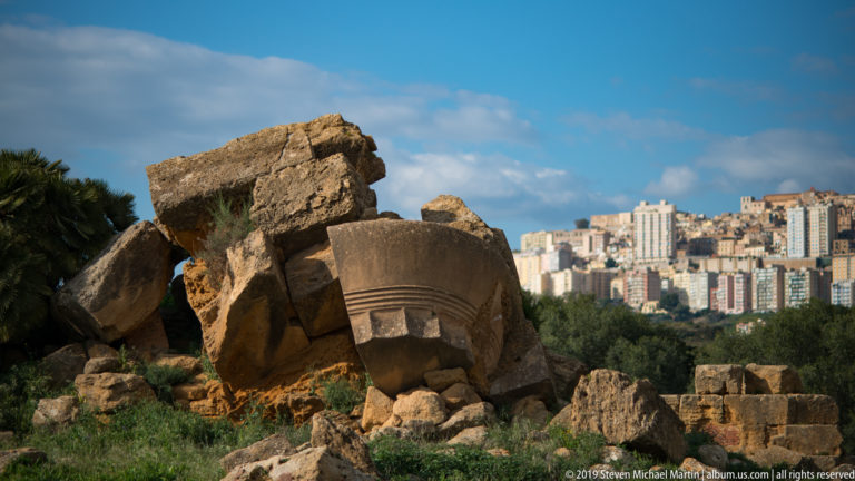 Massive fallen carved stone blocks in the foreground with modern Agrigento rising on a distant ridge under blue sky.
