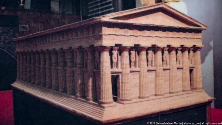 Scale model of a Greek temple with a pediment, deep colonnade, and large standing figures between exterior supports.
