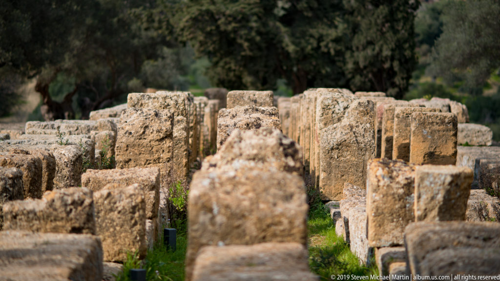 Closely spaced rows of weathered rectangular stone blocks with grass growing between them in an archaeological area.