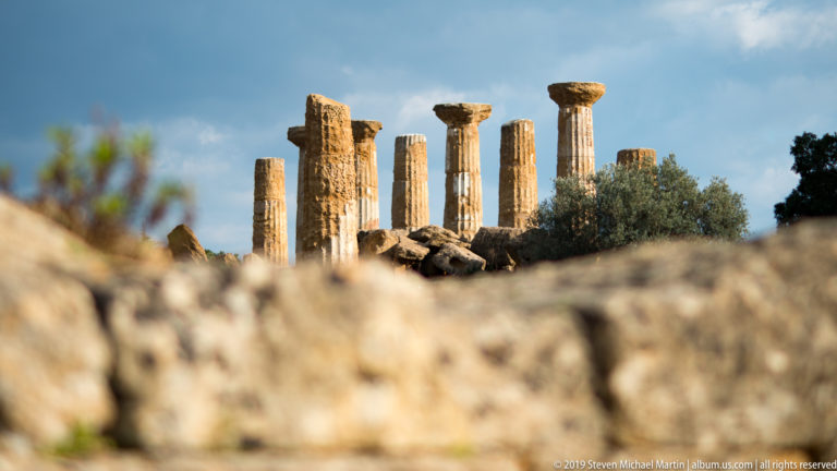 Row of weathered Doric columns and capitals rising beyond blurred stone ruins and an olive tree under blue sky.