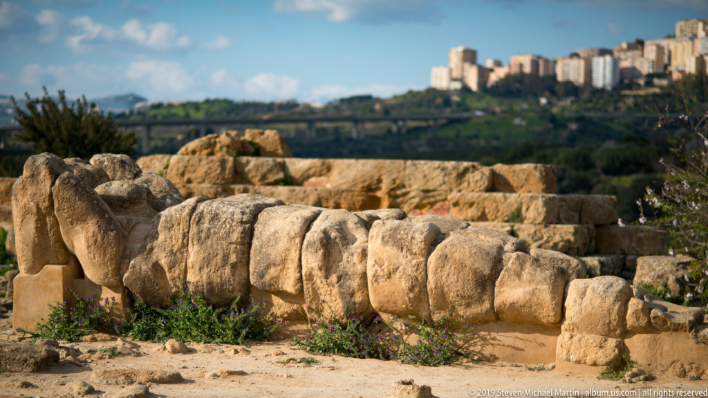 Side view of a colossal fallen stone figure among temple blocks, with modern Agrigento on the ridge in the background.