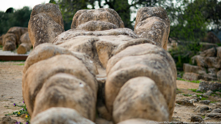 Foreshortened view from the feet of a colossal fallen stone male figure lying among archaeological ruins.