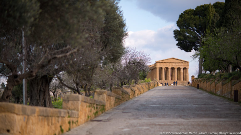 Tree-lined path and low stone walls converging on the distant sunlit Temple of Concordia with small visitors nearby.