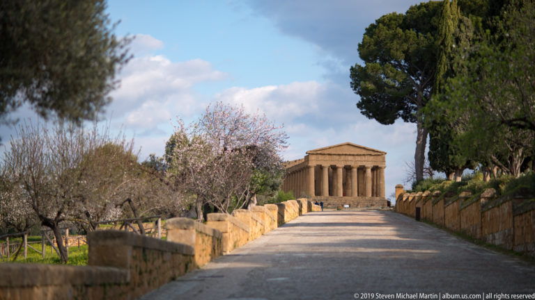Straight path lined by stone walls and trees leading toward the distant Temple of Concordia.