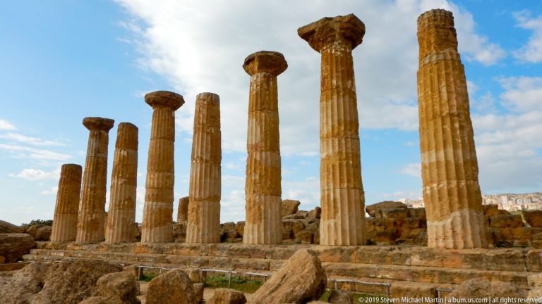 Row of tall weathered Doric columns of differing heights rising above broken stone ruins.