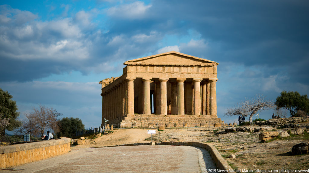 Temple of Concordia seen from the approach, with six Doric façade columns and triangular pediment under dark clouds.