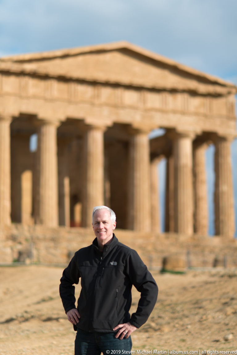 Man posing in front of the softly focused Temple of Concordia, with its pediment and Doric colonnade behind him.