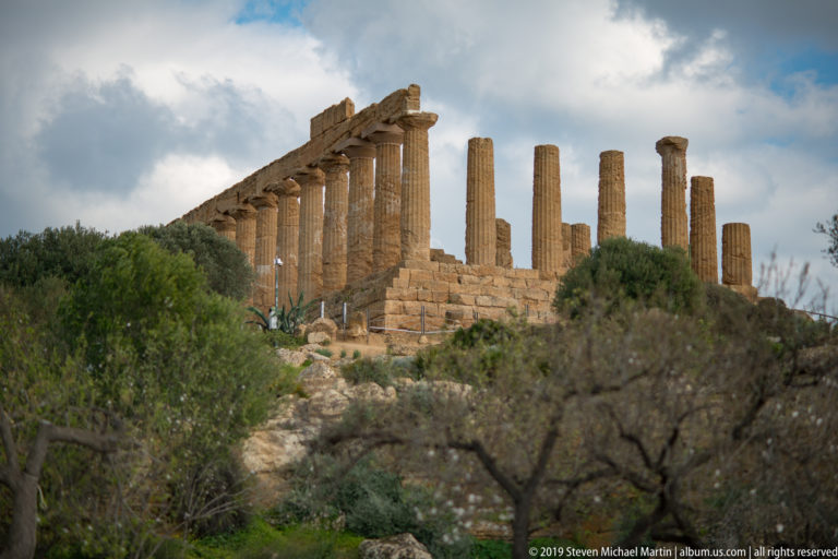 Ancient Doric temple columns rising above olive trees and shrubs on a rocky ridge beneath a cloudy sky.