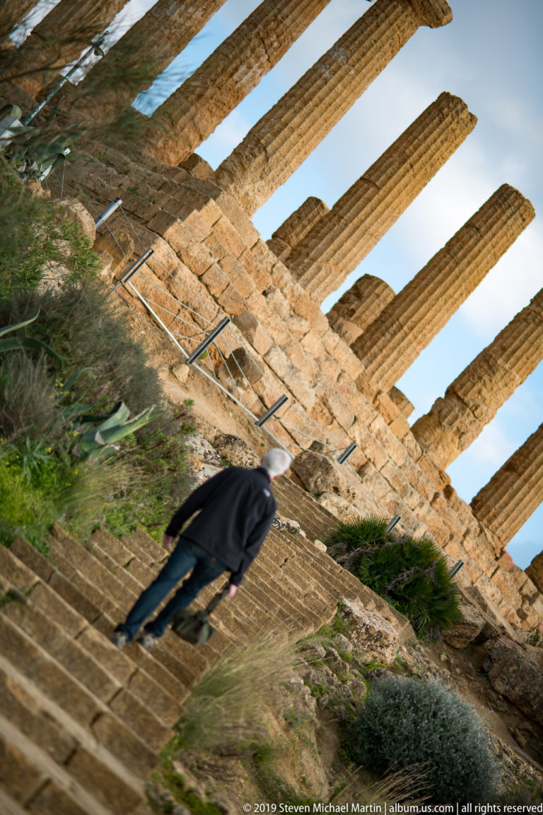 Angled upward view along ancient Doric columns, with stone steps, vegetation, and metal handrails below.