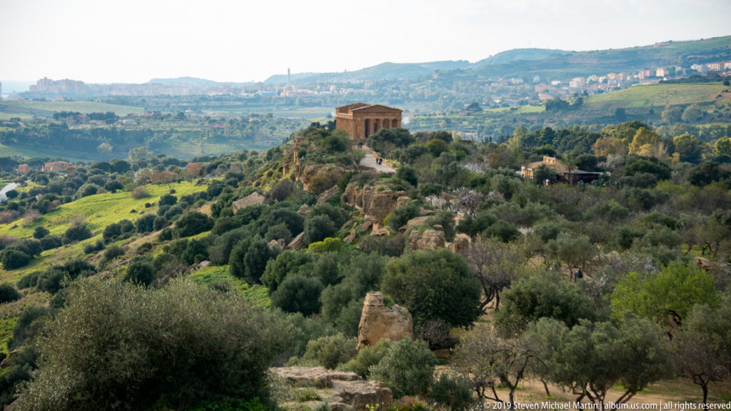 Distant Greek temple surrounded by olive trees and ruins, with the modern city of Agrigento on the ridge beyond.