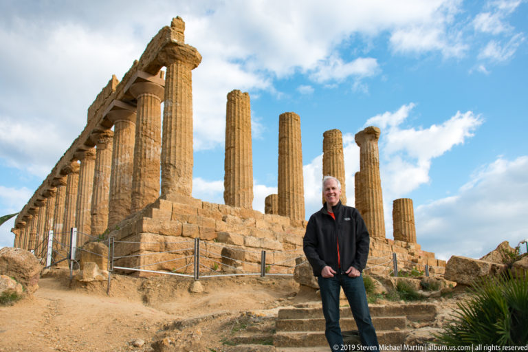 Man standing beside a long row of ancient Doric columns on a stepped stone platform under a cloudy sky.