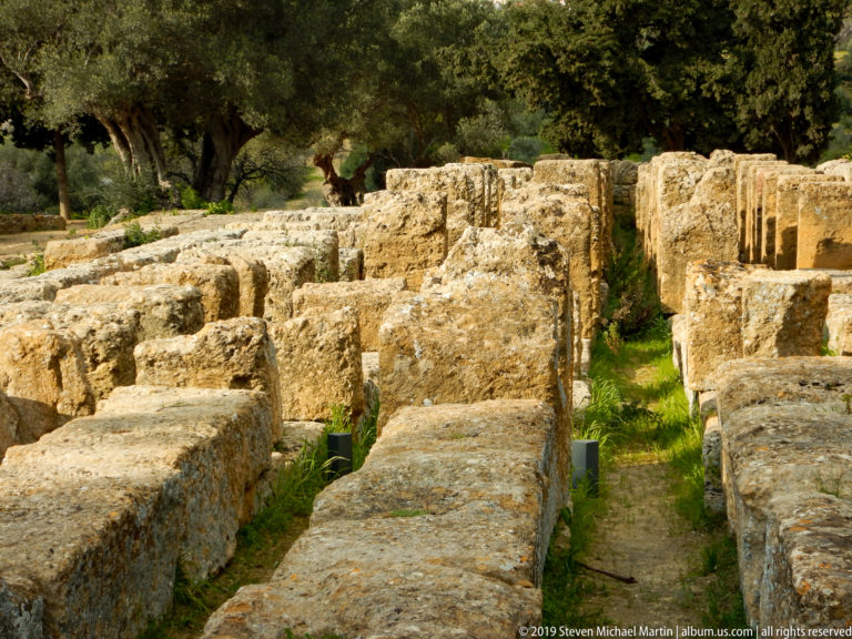Rows of massive weathered rectangular stone blocks separated by narrow grassy passages beneath olive trees.