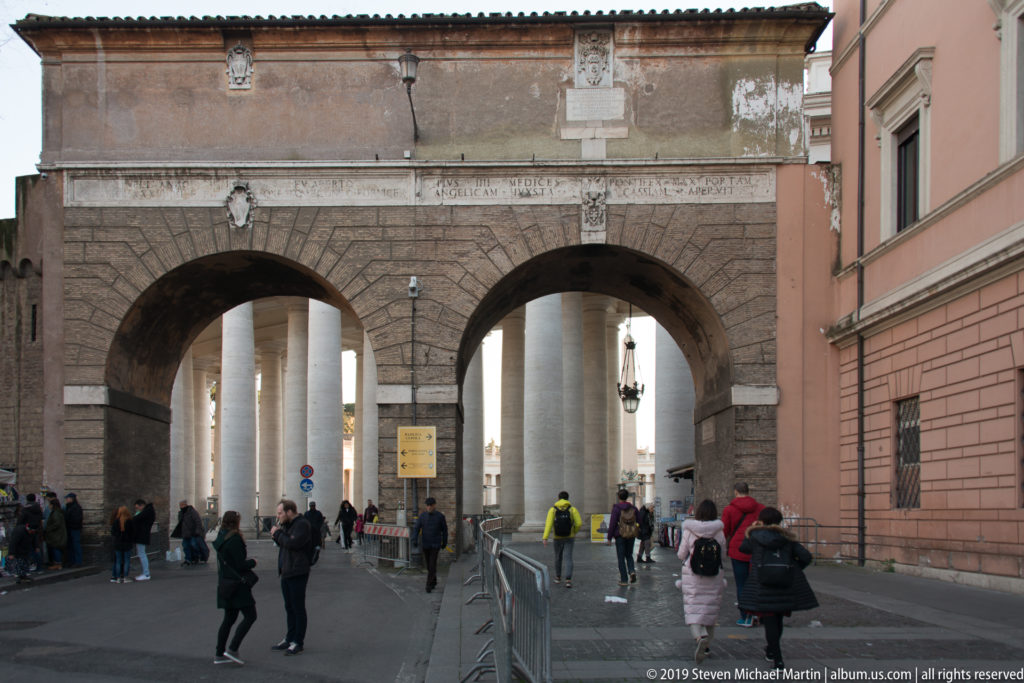 Twin arched masonry gateway with visitors and pedestrian barriers, opening toward the pale columns of Saint Peter’s Square.