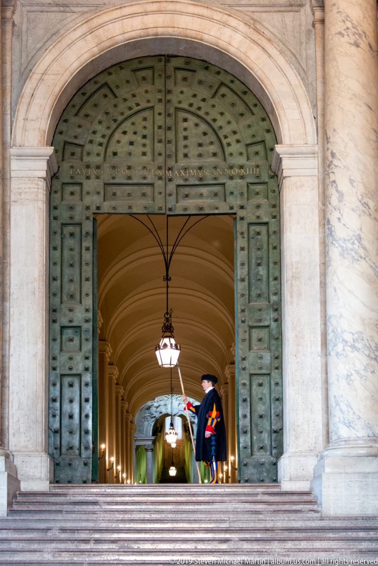 Open studded bronze doors beneath a marble arch lead into a vaulted passage where a Swiss Guard stands beside hanging lanterns.
