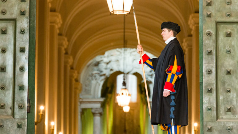 Pontifical Swiss Guard holding a polearm between open bronze doors, with vaulted passage and hanging lamps receding behind him.