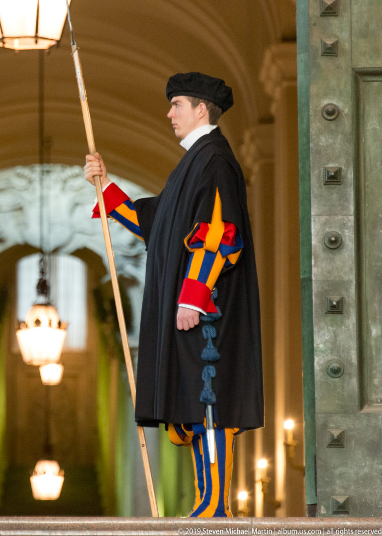 Pontifical Swiss Guard in blue, red, and yellow uniform and dark cloak, holding a polearm beside a large bronze doorway.