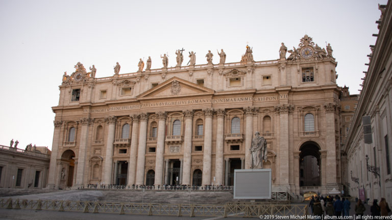 Wide exterior view of Saint Peter’s Basilica façade with giant columns, central balcony, attic inscription, and rooftop statues.