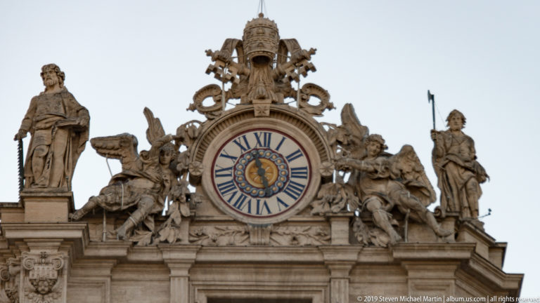 Large Roman-numeral clock on Saint Peter’s façade, surrounded by stone figures and elaborate heraldic and ornamental sculpture.