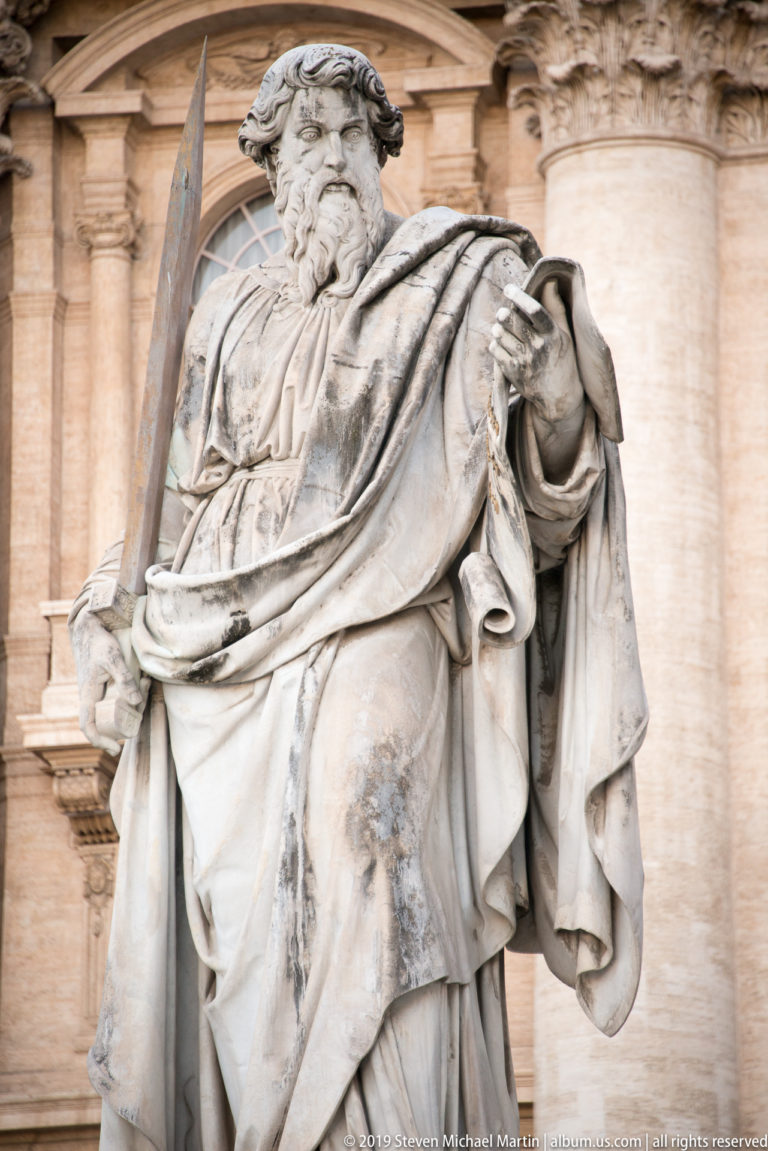 Colossal robed saint standing on the balustrade above the columns of Saint Peter’s Basilica façade.
