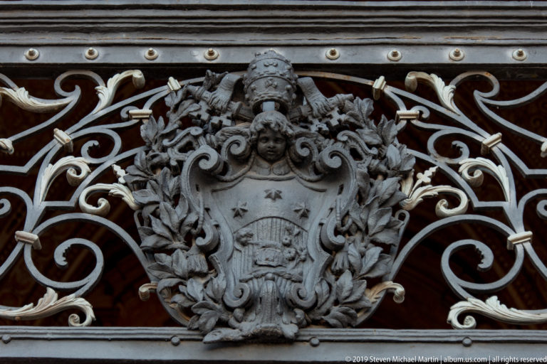 Papal coat of arms with tiara and crossed keys centered in ornate scrolling metalwork against a dark red background.