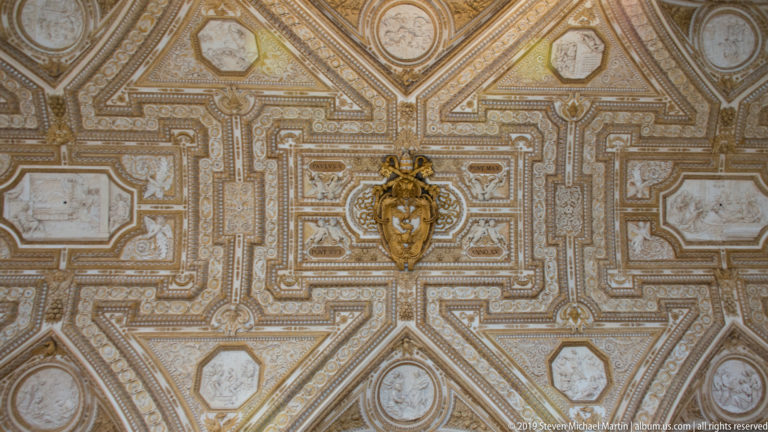 Ornate compartmented ceiling with gilded borders, relief medallions, rosettes, and a projecting papal heraldic emblem at the center.