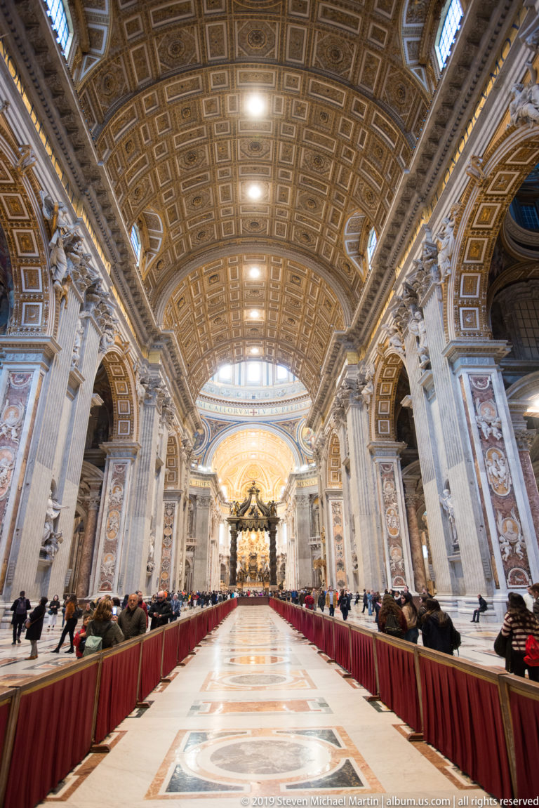 Centered low-angle view along the nave, showing patterned marble pavement, red barriers, visitors, giant piers, and gilded coffered ceiling.