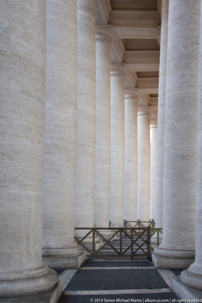 Close view between massive pale columns of Bernini’s colonnade, with a wooden barrier and repeated ceiling bays receding into the distance.