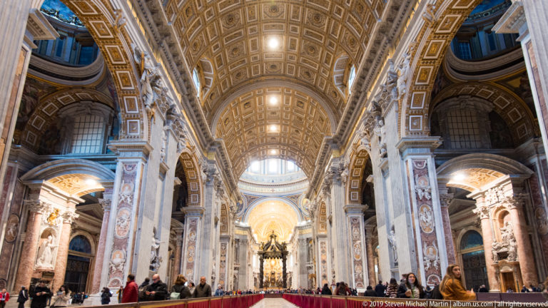 Long view down Saint Peter’s central nave beneath a gilded coffered vault, with massive piers, side arches, visitors, and red barriers.