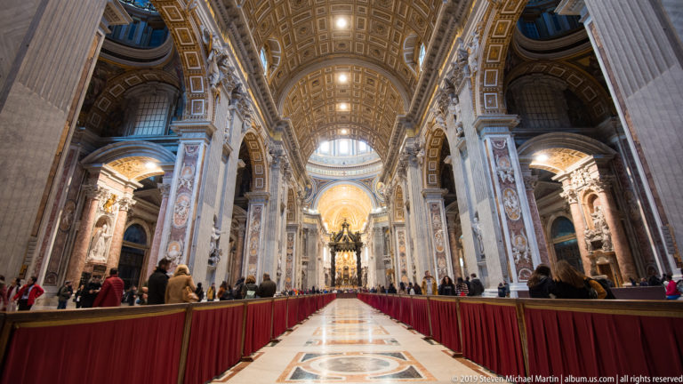 Vertical view down the nave toward the crossing, with gilded coffered vault, massive piers, side bays, visitors, and red barriers.