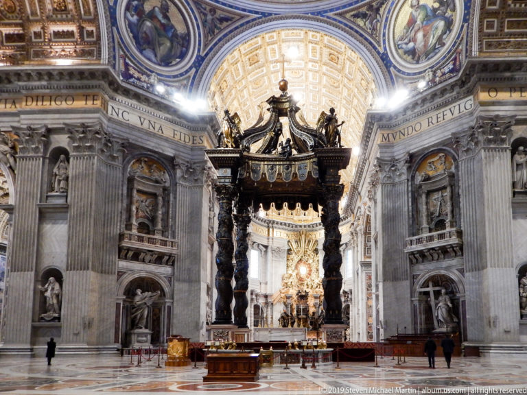 Bernini’s baldachin centered between massive stone piers, with niche statues, upper mosaics, and the bright apse beyond.