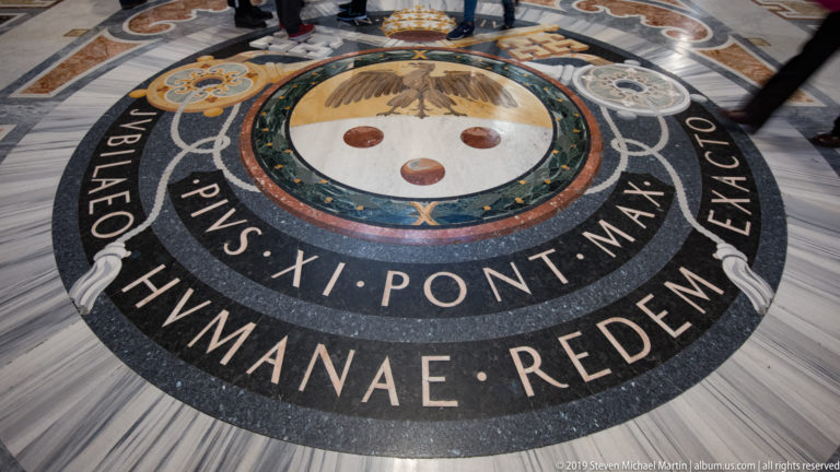 Circular marble floor inlay inscribed for Pius XI, with papal heraldry and concentric bands of green, black, cream, and red stone.