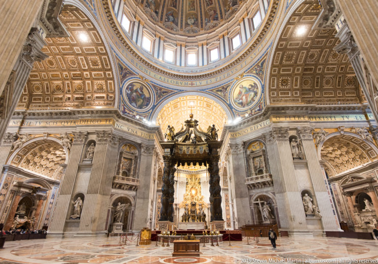 Wide crossing view with Bernini’s baldachin centered beneath the dome, surrounded by monumental piers, niches, mosaics, and inscriptions.
