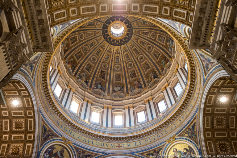 Upward view into Saint Peter’s great dome, with tall drum windows, gilded ribs, mosaic figures, and encircling inscriptions.