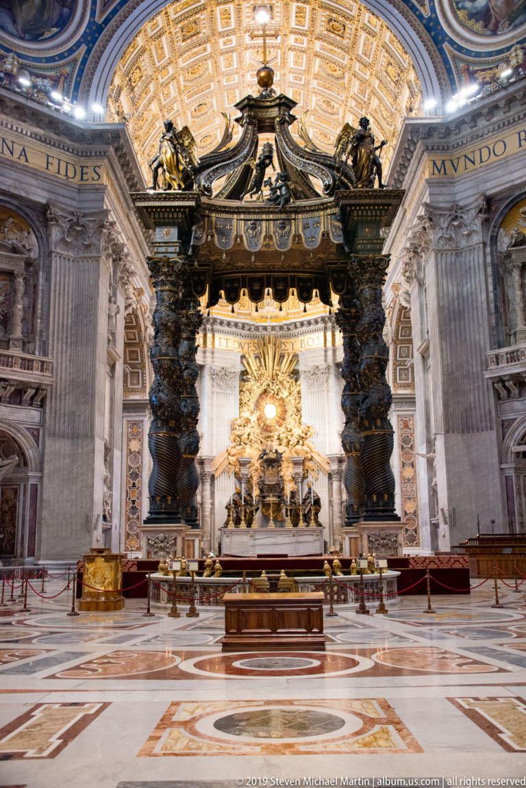 Wide interior view of Bernini’s four-column baldachin over the papal altar, aligned with the glowing Cathedra Petri beyond.