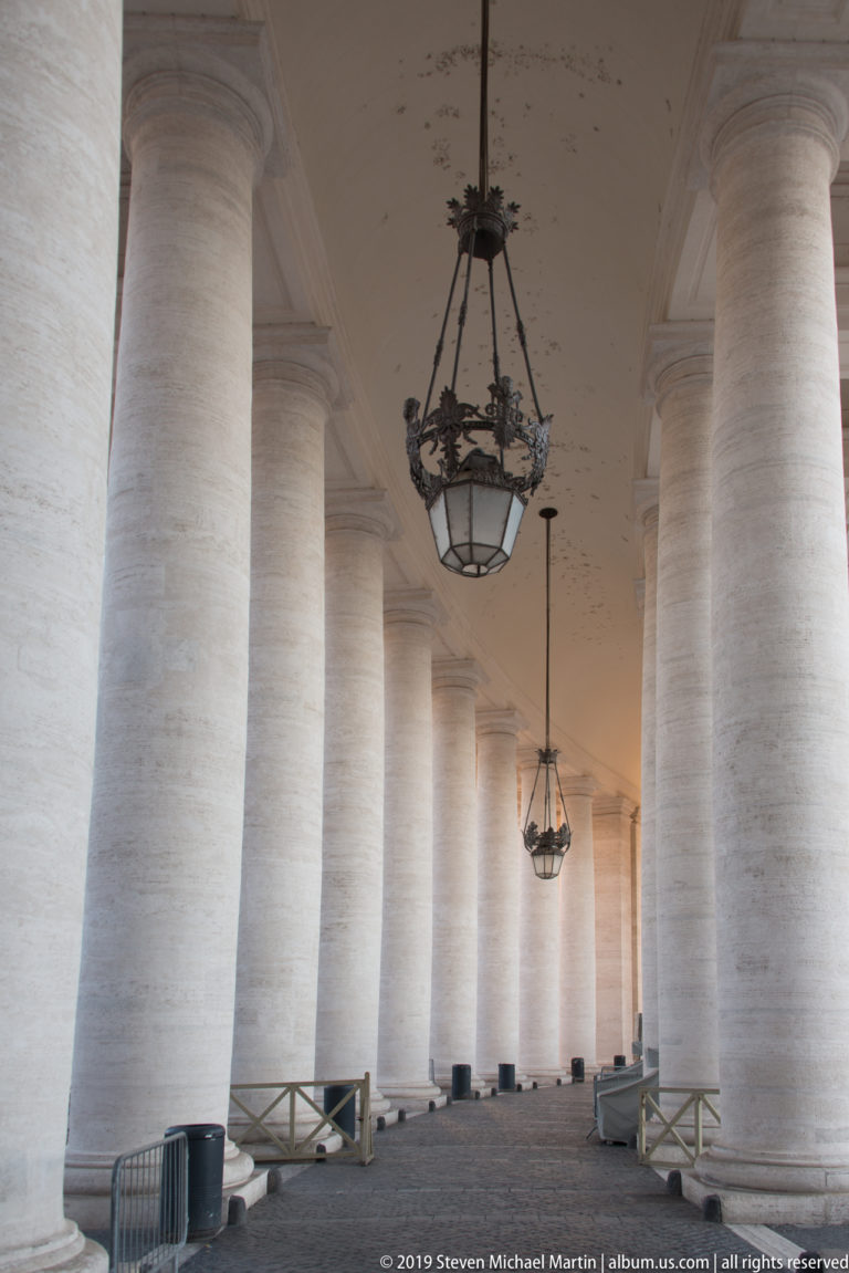 Covered passage through Bernini’s colonnade with repeated massive columns, hanging lanterns, pale paving, and low wooden barriers.