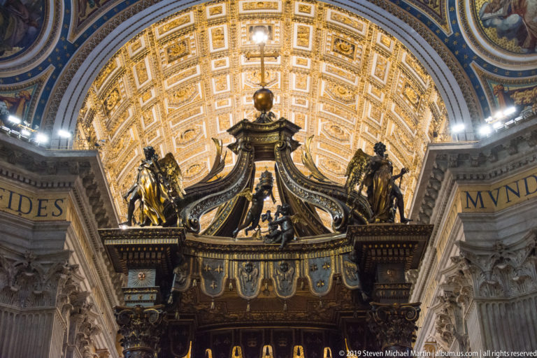 Upper crown of Bernini’s bronze baldachin with volutes, angels, gilded details, orb, and cross beneath a coffered vault.