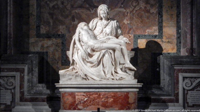 Michelangelo’s white marble Pietà on a reddish stone base against a dark polychrome marble wall inside Saint Peter’s Basilica.