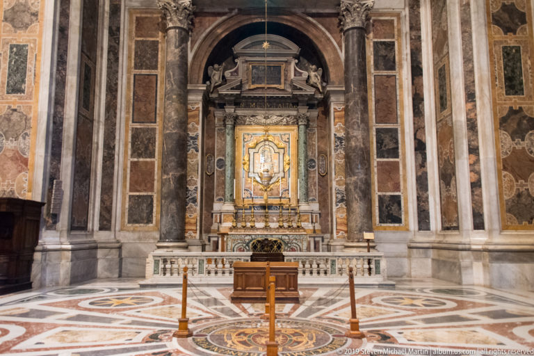 Frontal view of a chapel altar with gilded central object, paired marble columns, patterned wall panels, and geometric stone pavement.