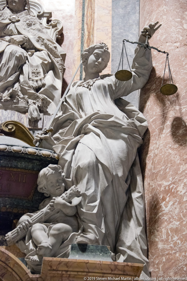 Reclining marble allegorical figure holding a balance scale beside a monument, viewed from below against reddish marble.
