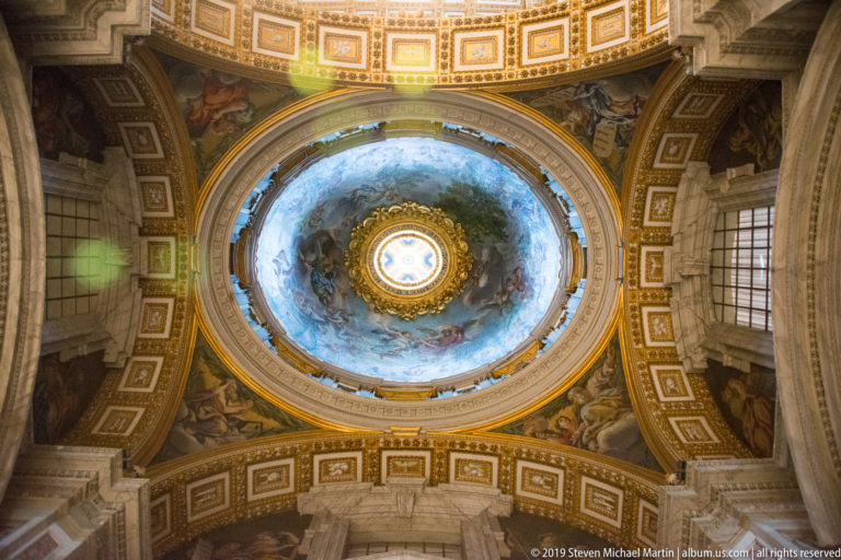 Upward view of a decorated cupola with a bright central oculus, blue painted figures, gilding, coffers, and surrounding architectural panels.