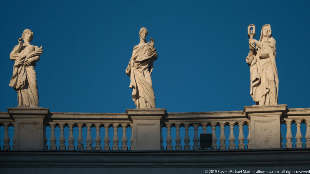 Three pale stone saint statues in varied poses stand on the balustrade above Bernini’s colonnade against a deep blue sky.