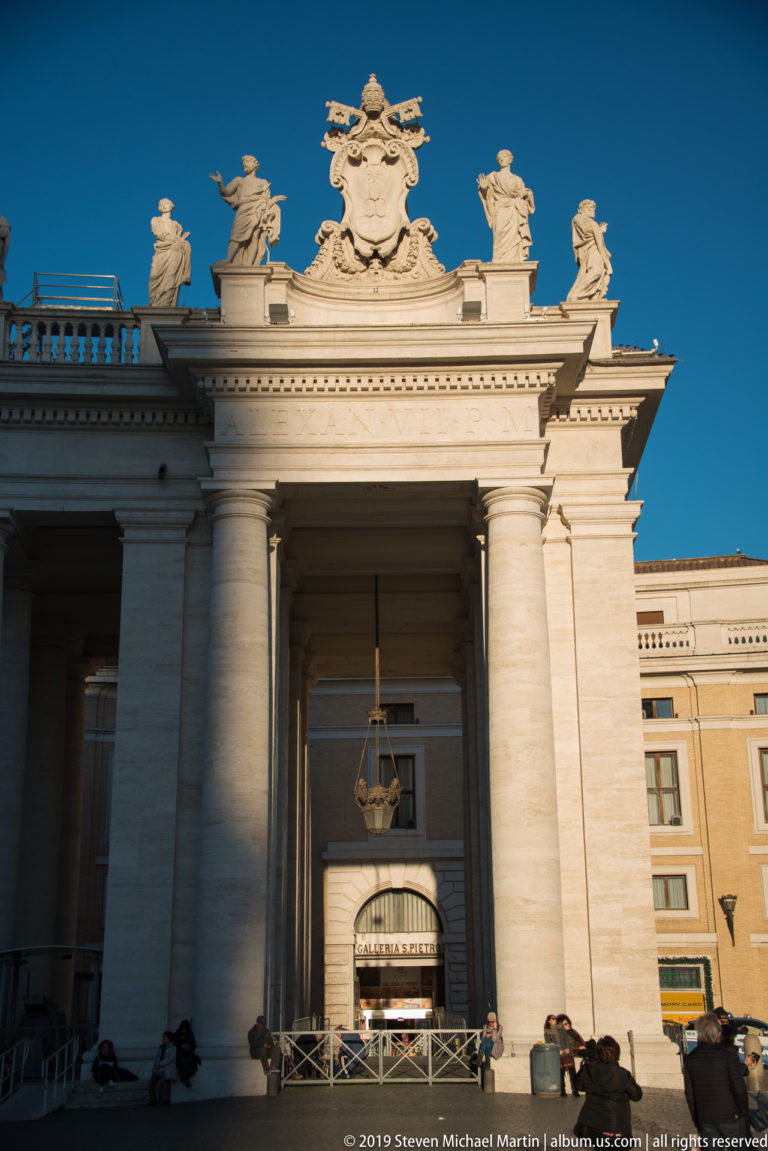 Tall entrance bay in Bernini’s colonnade with giant columns, papal coat of arms, rooftop statues, hanging lantern, and visitors.