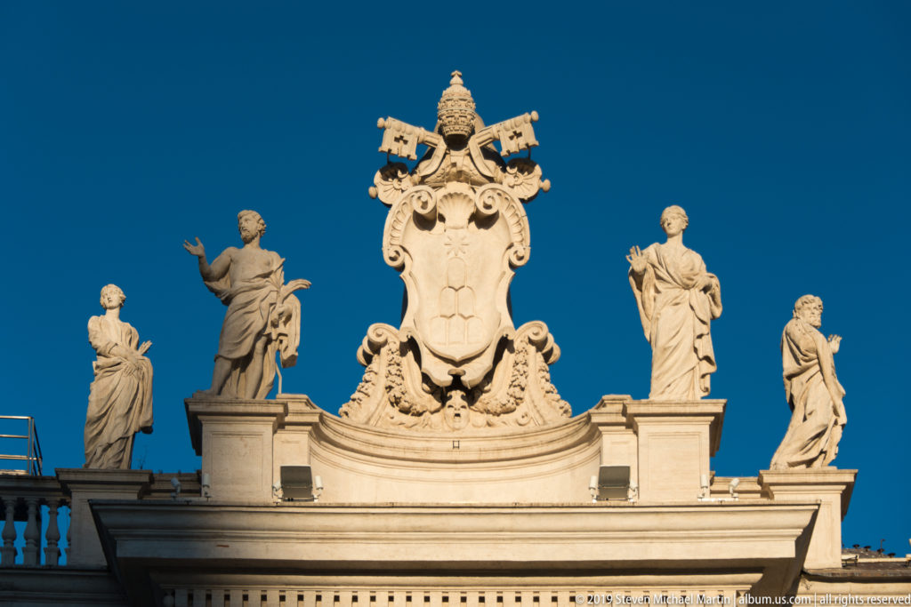 Large papal coat of arms with tiara and crossed keys centered between stone saint statues above the colonnade.