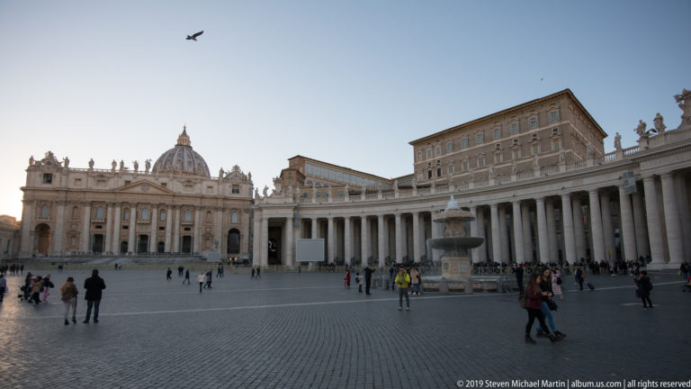 Wide view across Saint Peter’s Square toward the basilica, dome, curving colonnade, fountain, Apostolic Palace, and scattered visitors.