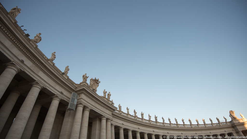 Low-angle view along Bernini’s colonnade with massive columns and a continuous row of saint statues silhouetted against blue sky.