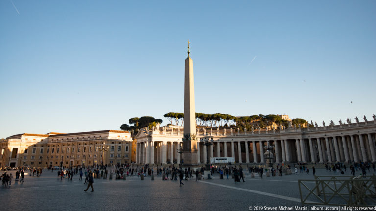 Vatican Obelisk rising in Saint Peter’s Square beside Bernini’s curved colonnade, rooftop statues, visitors, and distant umbrella pines.