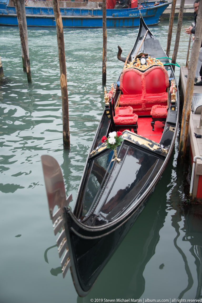 Black Venetian gondola moored beside wooden posts, with bright red upholstered passenger seats and a curved metal ferro at the prow.