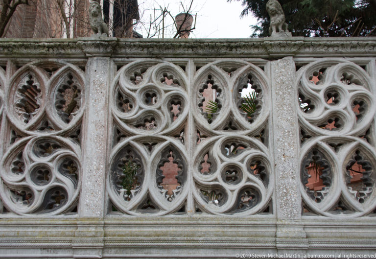 Close view of pale Gothic stone tracery with repeating quatrefoils, trefoils, pointed openings, and glimpses of greenery beyond.