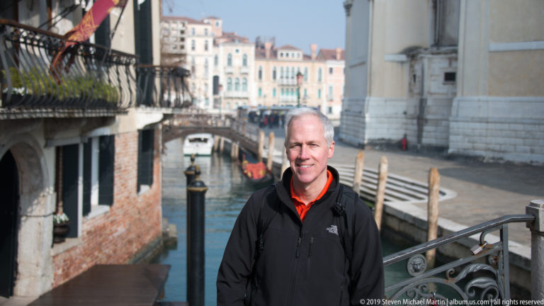 Man in a black jacket standing beside a Venetian canal with an arched bridge and mooring posts behind him.